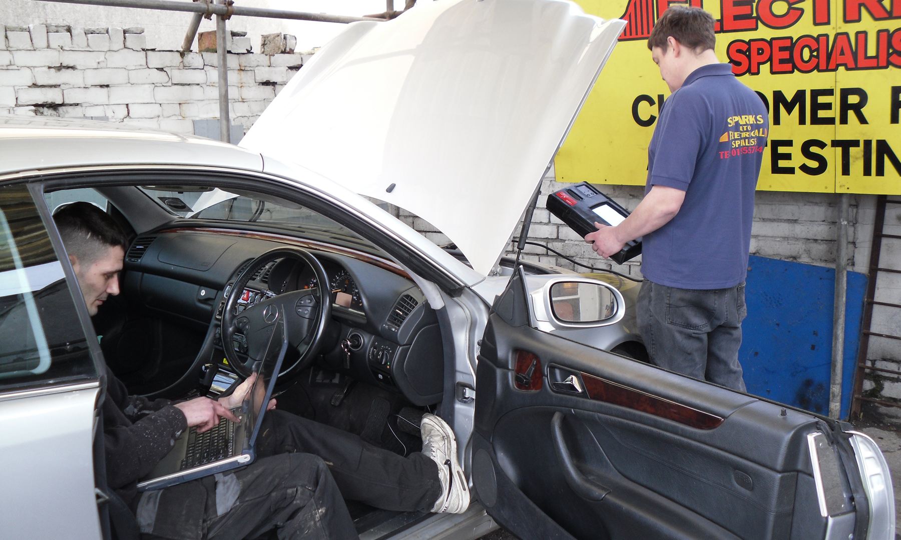 Two Sparks technicians diagnosing a silver Mercedes in the yard, one with a laptop in the driver's seat and one with a scan tool under the bonnet, the yellow Sparks sign behind